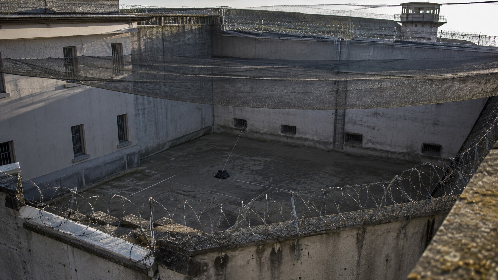 Cour de promenade d'une prison protégée par un filet anti-projection, un sac au sol relié à une ficelle